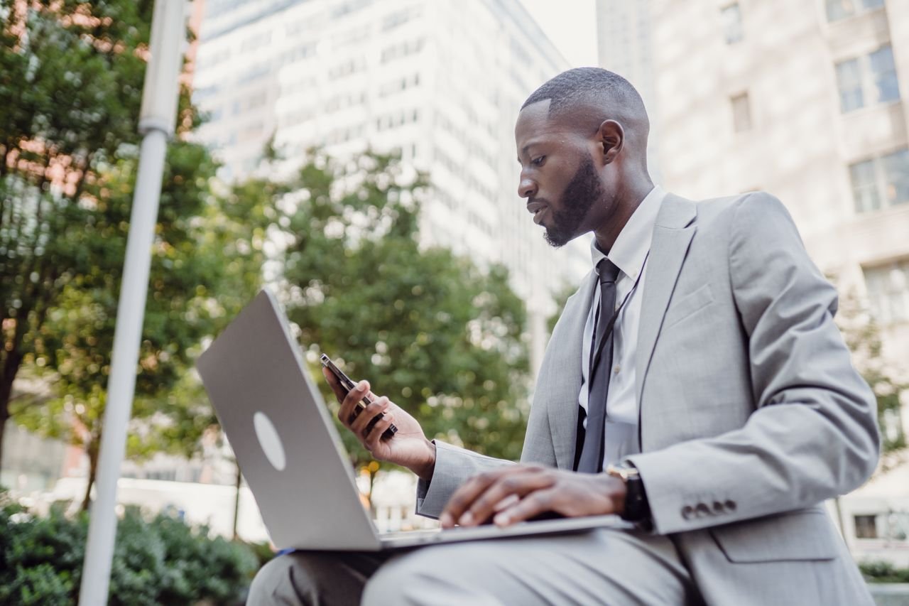 Businessman in a suit using a laptop and smartphone in an urban setting, blending technology with business.